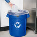 A hand pouring liquid from a plastic bottle into a blue Rubbermaid recycling bin.