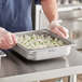 A person in a gloves and apron placing a Hatco half size stainless steel food pan full of food on a counter.