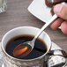 A hand holding a Chef & Sommelier stainless steel demitasse spoon over a cup of coffee.