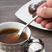 A hand holding a Chef & Sommelier stainless steel demitasse spoon over a cup of liquid.