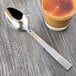 A Libbey stainless steel dessert spoon next to a glass of liquid on a wood table.