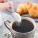 A hand holding a Libbey stainless steel demitasse spoon pouring sugar into a cup of coffee.