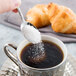A hand holding a Libbey stainless steel demitasse spoon pouring sugar into a cup of coffee.
