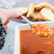 A hand holding a Libbey medium weight stainless steel bouillon spoon filled with soup over a bowl of soup.