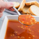 A finger holding a Libbey stainless steel bouillon spoon filled with soup over a bowl of soup.