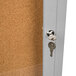 A satin anodized Aarco bulletin board cabinet with a cork board door and a key in the lock.