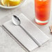 A Choice Windsor stainless steel iced tea spoon on a napkin next to a glass of orange liquid.