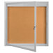 An Aarco indoor bulletin board cabinet with cork board and white frame with door open.