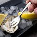 A hand holding a Chef & Sommelier stainless steel oyster fork over an oyster shell with a lemon wedge on ice.