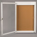 A white frame with a cork board inside and a glass door with a white frame and lock.
