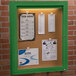 An Aarco green indoor lighted bulletin board cabinet with cork board inside and a green frame.