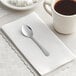 A Choice Windsor stainless steel demitasse spoon on a white napkin next to a cup of coffee.