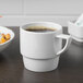 A white Schonwald stackable mug filled with brown liquid on a table with a bowl of food.