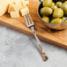 A Master's Gauge stainless steel cocktail fork on a cutting board with olives and cheese.