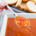 A finger holding a Reserve by Libbey stainless steel bouillon spoon filled with soup over a bowl of soup.