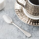 A silver Reserve by Libbey stainless steel demitasse spoon on a white plate with a silver cup and saucer.