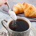 A hand holding a Reserve by Libbey stainless steel demitasse spoon pouring sugar into a cup of coffee.