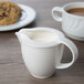 A white Villeroy & Boch porcelain creamer with a handle on a table with a cup of coffee and a cookie.