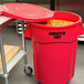 A red Rubbermaid BRUTE trash can with a lid open containing food in a school kitchen.