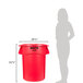A woman standing next to a Rubbermaid red round trash can with a lid.