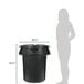A woman standing next to a black Rubbermaid BRUTE trash can with a lid.