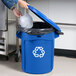 A person putting a plastic container into a blue Rubbermaid recycling bin with a blue lid.