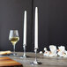 Two white Sterno taper candles in silver holders on a table with a glass of wine.