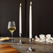 Two Sterno white taper candles on a wooden table with a glass of wine.
