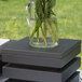 A Vollrath black wooden display platter with a glass jar of leaves in water on top of it.