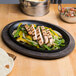A plate of grilled chicken, peppers, and tortillas on a Lodge oval cast iron fajita skillet with a wood underliner.