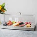 A Vollrath stainless steel cooling plate with a clear lid on a counter with cheese, grapes and a flower.