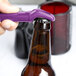 A hand holding a purple Franmara Traveler's Grape plastic bottle opener opening a brown bottle.