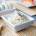 A white square ramekin filled with rice and sushi.