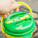 A hand holding a green and yellow 64 oz. lemonade bucket jug with a lid.