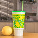 A white cylinder with a 32 oz. Lemonade Economy Car Cup with a green lid and a lemon.