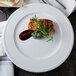 A Libbey alpine white porcelain plate with salmon, sauce, and herbs on a table.