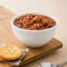 A Libbey alpine white porcelain bowl of chili with crackers and a spoon on a wooden surface.