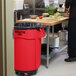 A man in a chef's outfit standing in front of a red Rubbermaid trash can in a school kitchen.