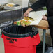 A person in a black chef's outfit putting food into a Rubbermaid red round trash can.