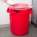 A hand holding a white plastic bag in a red Rubbermaid commercial trash can.