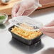 A gloved hand filling a black rectangular plastic container with macaroni and cheese.