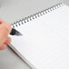 A hand holding a pen writes on a white wirebound spiral notebook.