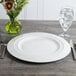 A white Bon Chef porcelain charger plate on a table with a glass of water and a vase of flowers.