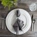 A Bon Chef white porcelain charger plate with silverware and a napkin on a table.