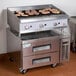 A Cooking Performance Group gas charbroiler on a stainless steel chef station counter with meat cooking on it.