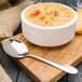 A Walco stainless steel bouillon spoon in a bowl of soup with vegetables on a wood board.
