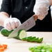 A person wearing gloves uses a Mercer Culinary Praxis chef's knife to slice a cucumber on a counter.