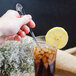 A hand holding a Walco stainless steel iced tea spoon over a glass of ice tea with a lemon slice.