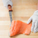 A person wearing gloves uses a Mercer Culinary Praxis stiff boning knife to cut a piece of salmon.