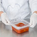 A person in gloves using a Carlisle clear polycarbonate hinged lid on a container of red sauce.